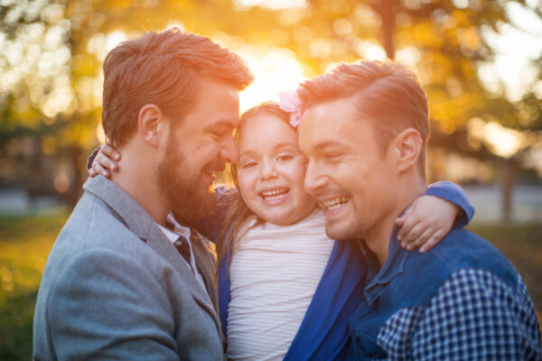 Young gay parents with their daughter having fun in park. Parents holding girl in arms. Enjoying in beautiful sunset. Caucasian ethnicity.